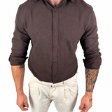 A man wearing a dark brown long-sleeve linen button-up shirt and off-white linen trousers, shown from the neck down against a plain white background.