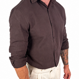 A man wearing a dark brown long-sleeve button-up shirt and off-white trousers, shown from the neck down against a plain white background.