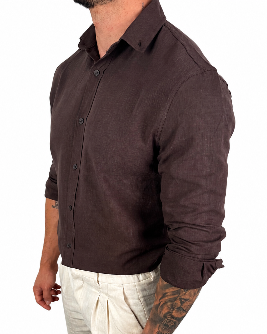 A man wearing a dark brown long-sleeve linen button-down shirt and off-white linen trousers, shown from a side profile against a white background.