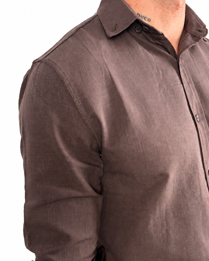 A man wearing a dark brown linen button-down shirt with rolled-up sleeves and off-white trousers, shown from the chest down against a white background.