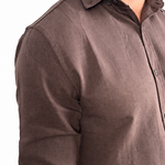 A man wearing a dark brown linen button-down shirt with rolled-up sleeves and off-white trousers, shown from the chest down against a white background.