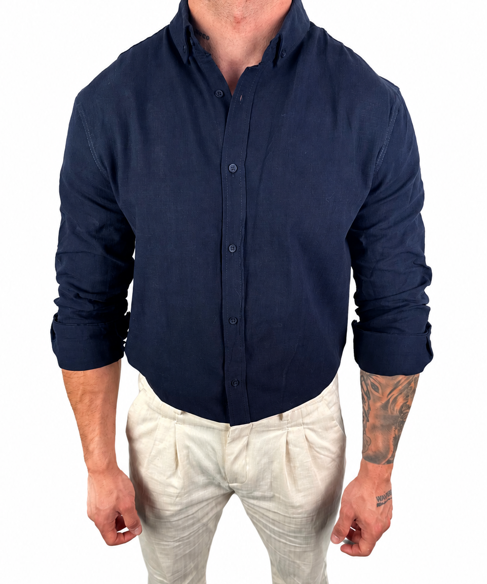 A man wearing a dark navy blue linen button-down shirt and off-white linen trousers, standing against a solid white background.