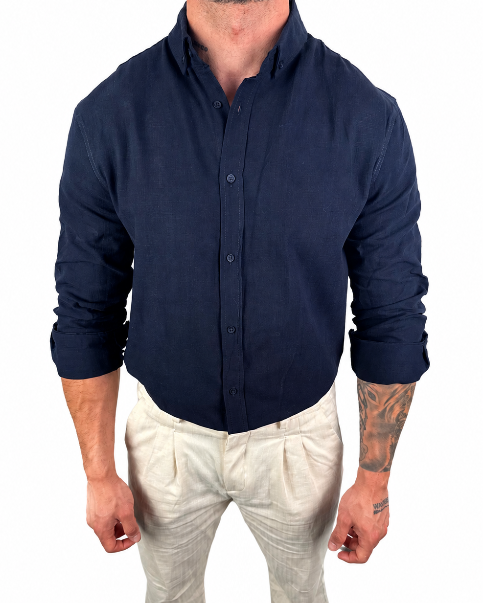 A man wearing a dark navy blue linen button-down shirt and off-white linen trousers, standing against a solid white background.