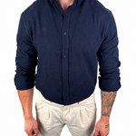 A man wearing a dark navy blue linen button-down shirt and off-white linen trousers, standing against a solid white background.