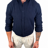 A man wearing a dark navy blue linen button-down shirt and off-white linen trousers, standing against a solid white background.
