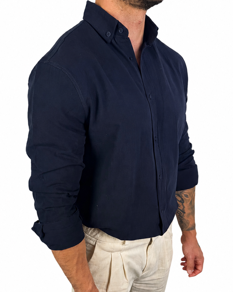 A man wearing a dark navy blue long-sleeve button-down shirt and light beige trousers, shown from the waist up against a plain white background.