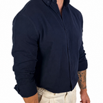 A man wearing a dark navy blue long-sleeve button-down shirt and light beige trousers, shown from the waist up against a plain white background.