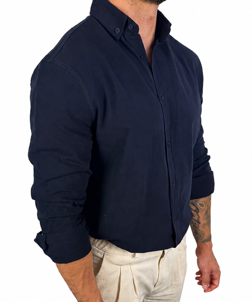 A man wearing a dark navy blue long-sleeve button-down shirt and light beige trousers, shown from the waist up against a plain white background.