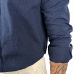 Close-up of a person wearing a navy blue long-sleeve linen shirt with rolled cuffs, paired with light beige trousers, against a white background.
