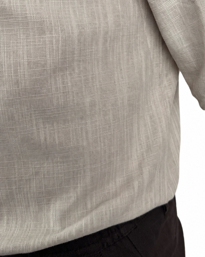 Close-up of a man wearing a light beige linen shirt with rolled-up sleeves and dark trousers, against a plain white background.