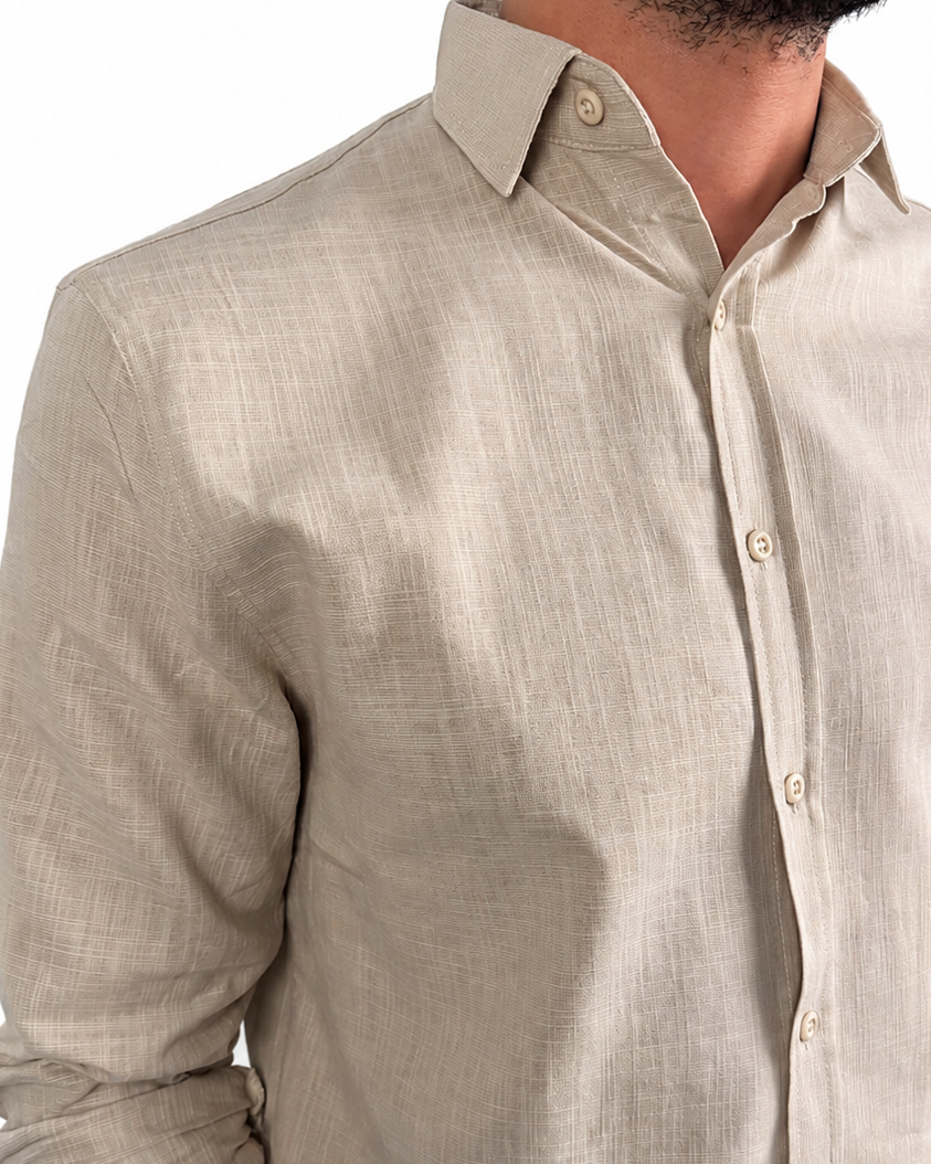 A man wearing a light beige linen button-down shirt with rolled-up sleeves and dark brown trousers, shown from the chest down against a white background.
