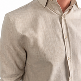 A man wearing a light beige linen button-down shirt with rolled-up sleeves and dark brown trousers, shown from the chest down against a white background.