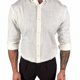 A man wearing a light beige linen long-sleeve button-down shirt with rolled-up sleeves and black trousers, shown from the neck down against a plain white background.