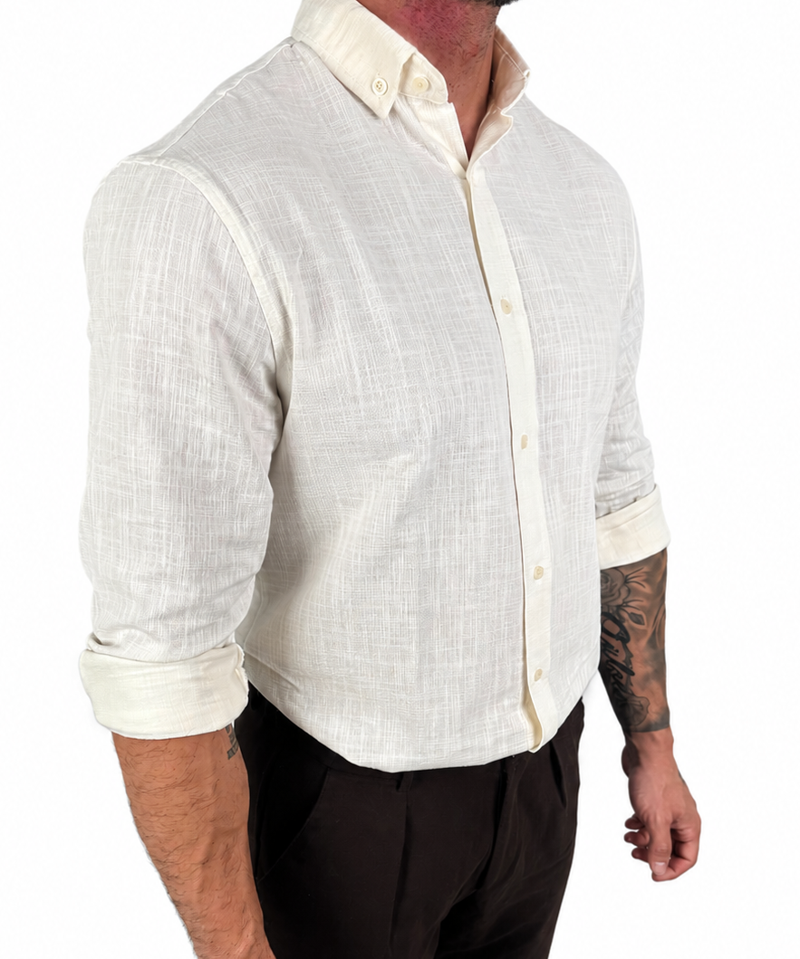 A man wearing a light cream-colored linen button-down shirt with rolled-up sleeves and dark brown trousers, shown from the waist up against a plain white background.