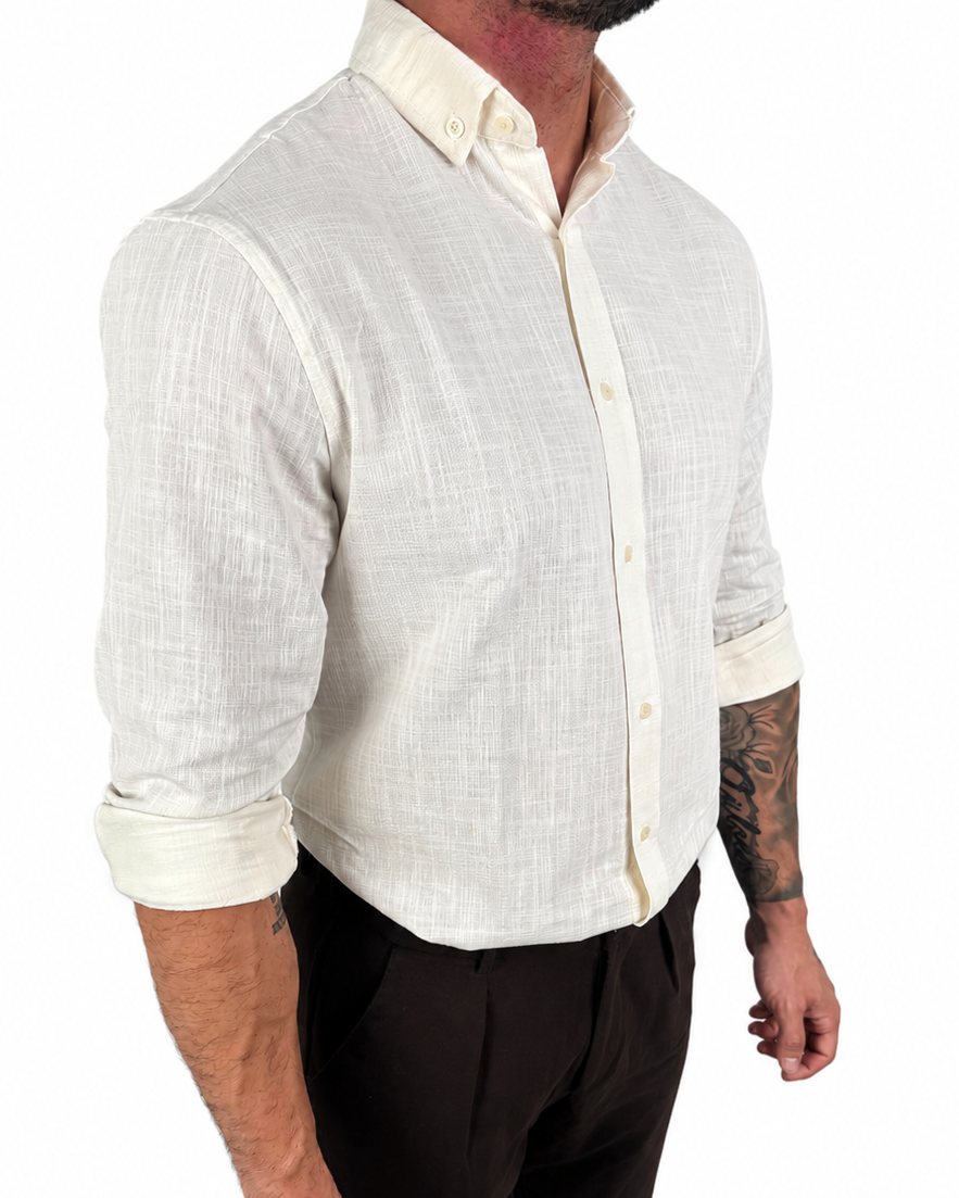 A man wearing a light cream-colored linen button-down shirt with rolled-up sleeves and dark brown trousers, shown from the waist up against a plain white background.