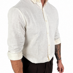 A man wearing a light cream-colored linen button-down shirt with rolled-up sleeves and dark brown trousers, shown from the waist up against a plain white background.