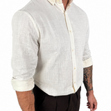A man wearing a light cream-colored linen button-down shirt with rolled-up sleeves and dark brown trousers, shown from the waist up against a plain white background.
