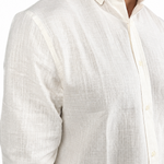 A close-up of a man wearing a textured off-white linen button-down shirt with rolled-up sleeves, paired with dark trousers.