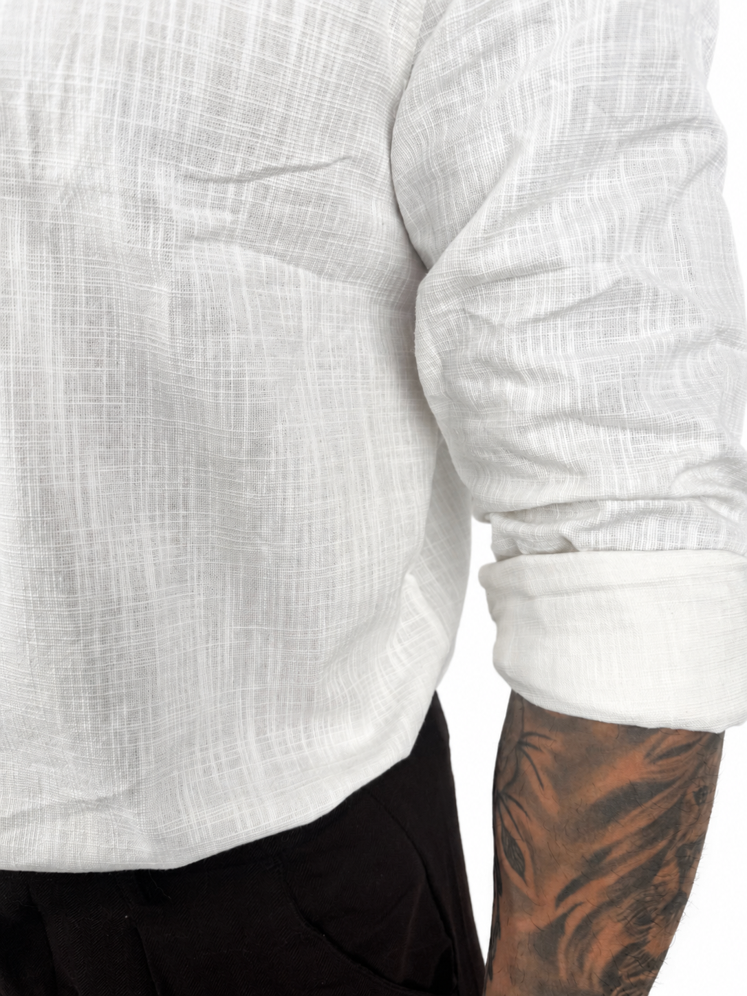 Close-up of a person wearing a white textured long-sleeve shirt with rolled sleeves and black pants, showing a tattooed arm against a plain white background.