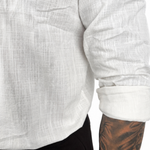 Close-up of a person wearing a white textured long-sleeve shirt with rolled sleeves and black pants, showing a tattooed arm against a plain white background.