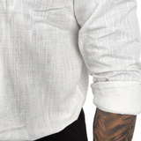 Close-up of a person wearing a white textured long-sleeve shirt with rolled sleeves and black pants, showing a tattooed arm against a plain white background.