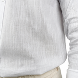 Close-up of a person wearing a white long-sleeve linen shirt tucked into light beige trousers, with a tattooed arm visible at the side.