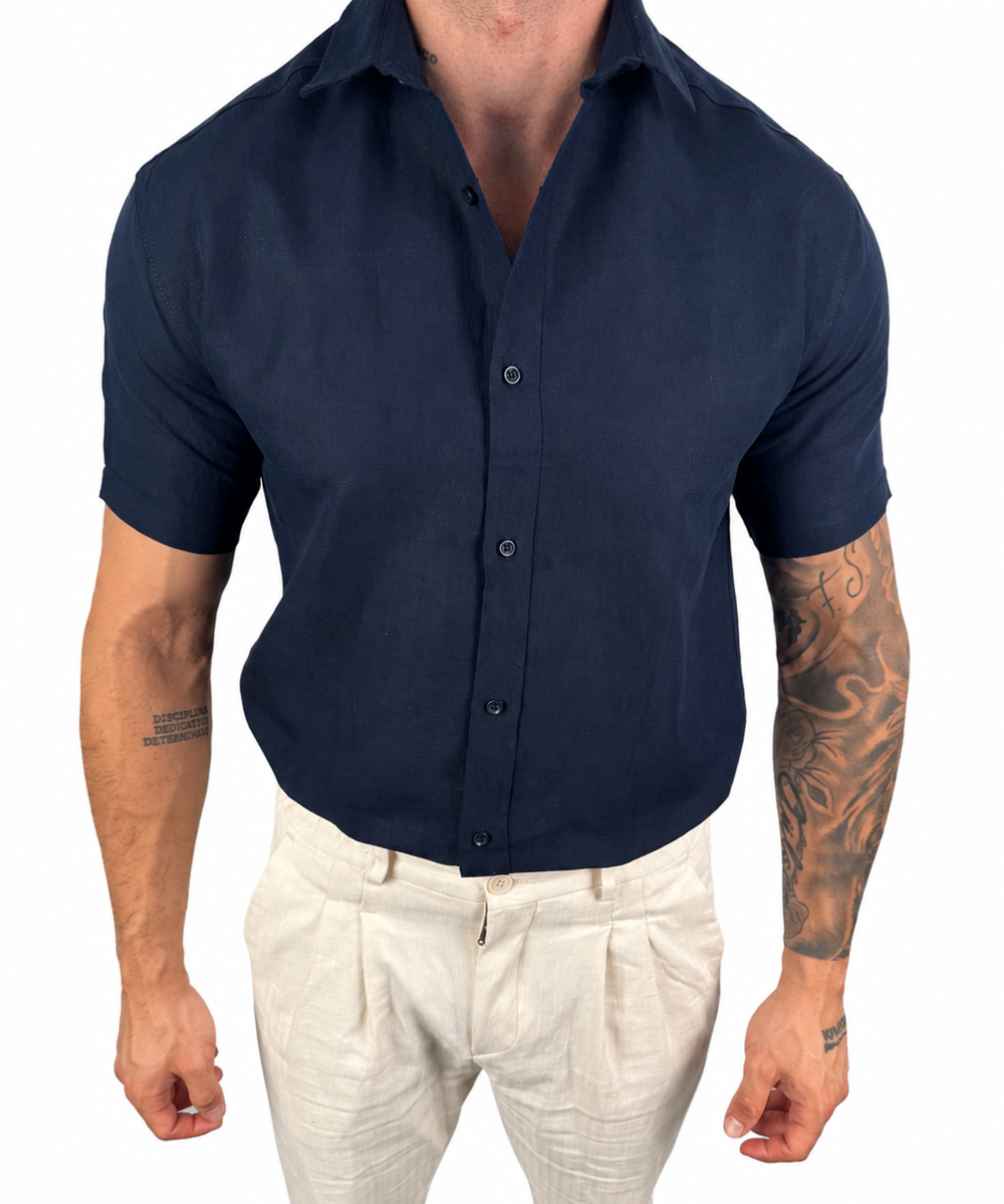A man wearing a dark navy blue short-sleeve button-down shirt tucked into off-white trousers, shown from the neck down against a plain white background.