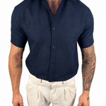 A man wearing a dark navy blue short-sleeve button-down shirt tucked into off-white trousers, shown from the neck down against a plain white background.