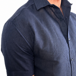 A man wearing a dark navy blue short-sleeve linen button-down shirt, shown from the chest down against a plain white background.