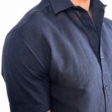 A man wearing a dark navy blue short-sleeve linen button-down shirt, shown from the chest down against a plain white background.