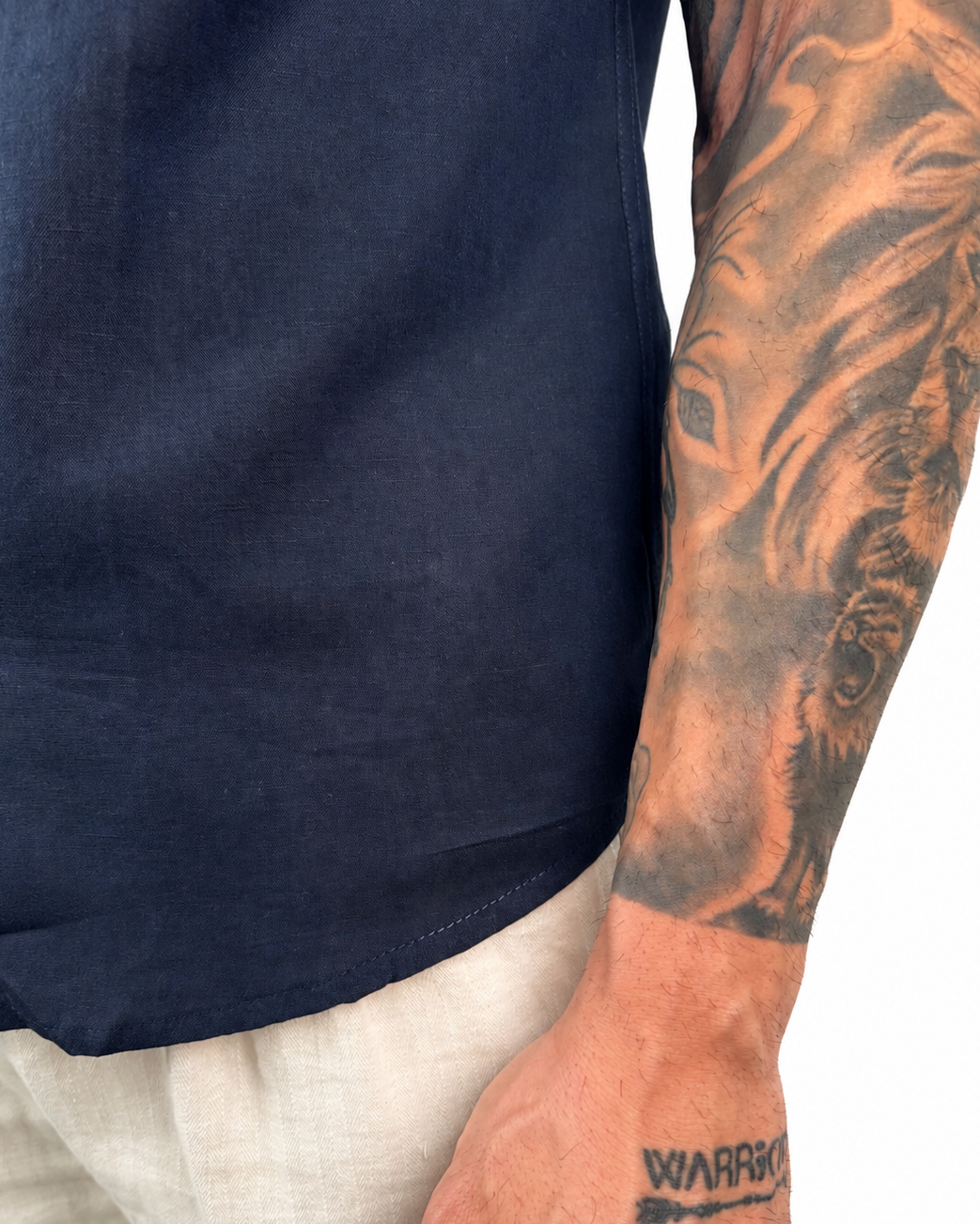 Close-up of a person wearing a navy blue short-sleeve shirt and light beige linen trousers, with a detailed tattoo on the right arm and a small 'WARPO' tattoo on the wrist.