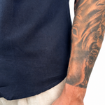 Close-up of a person wearing a navy blue short-sleeve shirt and light beige linen trousers, with a detailed tattoo on the right arm and a small 'WARPO' tattoo on the wrist.