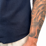 Close-up of a person wearing a navy blue short-sleeve shirt and light beige linen trousers, with a detailed tattoo on the right arm and a small 'WARPO' tattoo on the wrist.