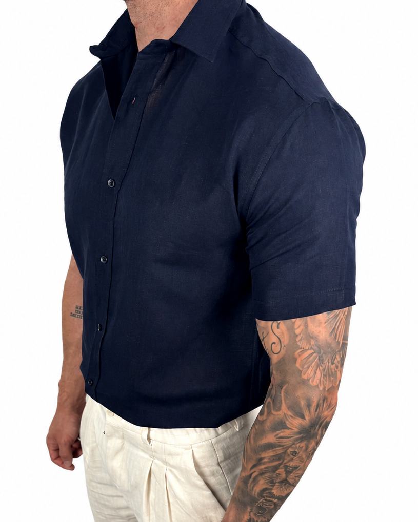 A muscular man wearing a dark navy blue short-sleeve button-up shirt and off-white linen trousers, shown from a side profile against a white background.