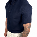 A muscular man wearing a dark navy blue short-sleeve button-up shirt and off-white linen trousers, shown from a side profile against a white background.