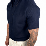A muscular man wearing a dark navy blue short-sleeve button-up shirt and off-white linen trousers, shown from a side profile against a white background.