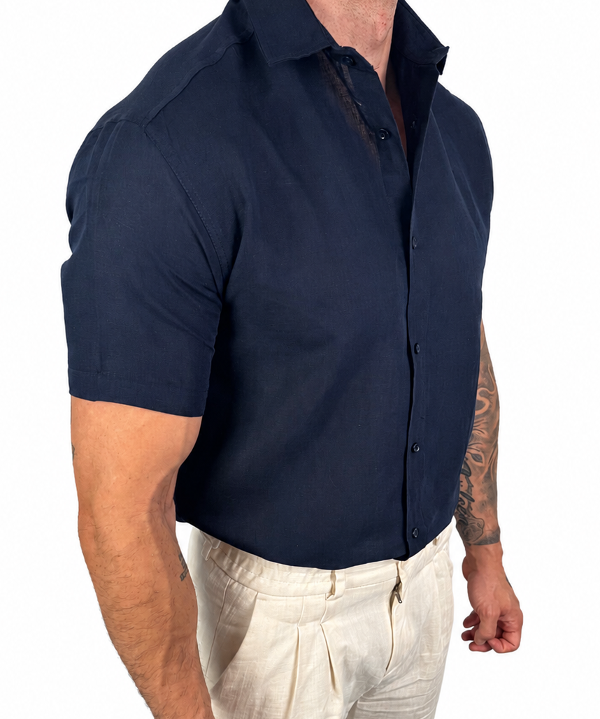 A muscular man wearing a navy blue short-sleeve button-down shirt and off-white linen trousers, shown from the waist down against a plain white background.
