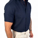 A muscular man wearing a navy blue short-sleeve button-down shirt and off-white linen trousers, shown from the waist down against a plain white background.