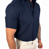 A muscular man wearing a navy blue short-sleeve button-down shirt and off-white linen trousers, shown from the waist down against a plain white background.