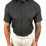 A man wearing a dark charcoal gray short-sleeve button-down shirt tucked into light beige linen trousers, shown from the neck down against a plain white background.