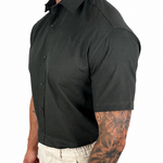 A man wearing a dark charcoal gray short-sleeve button-down shirt and off-white trousers, shown from a side profile against a plain white background.