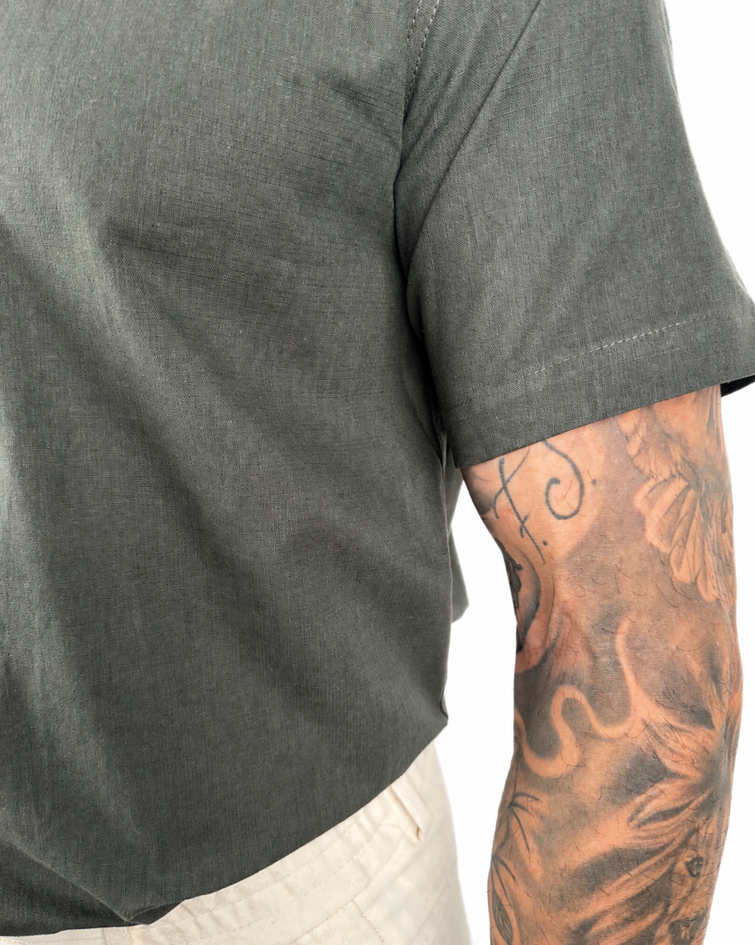 Close-up of a person wearing a olive green short-sleeve t-shirt and off-white pants, showing a detailed tattooed arm against a white background.