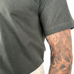 Close-up of a person wearing a olive green short-sleeve t-shirt and off-white pants, showing a detailed tattooed arm against a white background.