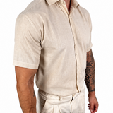 A man wearing a light beige short-sleeve linen button-up shirt and matching beige trousers, shown from the chest down against a plain white background.