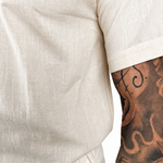 Close-up of a person wearing a short-sleeve cream-colored linen shirt, showing the textured fabric and a tattooed arm.