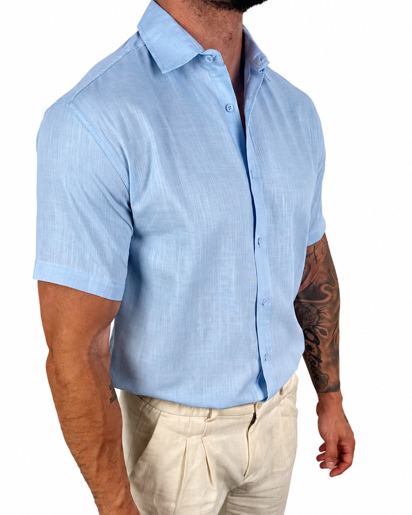 A man wearing a light blue short-sleeve linen button-down shirt and beige trousers, shown from the waist down against a plain white background.