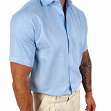 A man wearing a light blue short-sleeve linen button-down shirt and beige trousers, shown from the waist down against a plain white background.