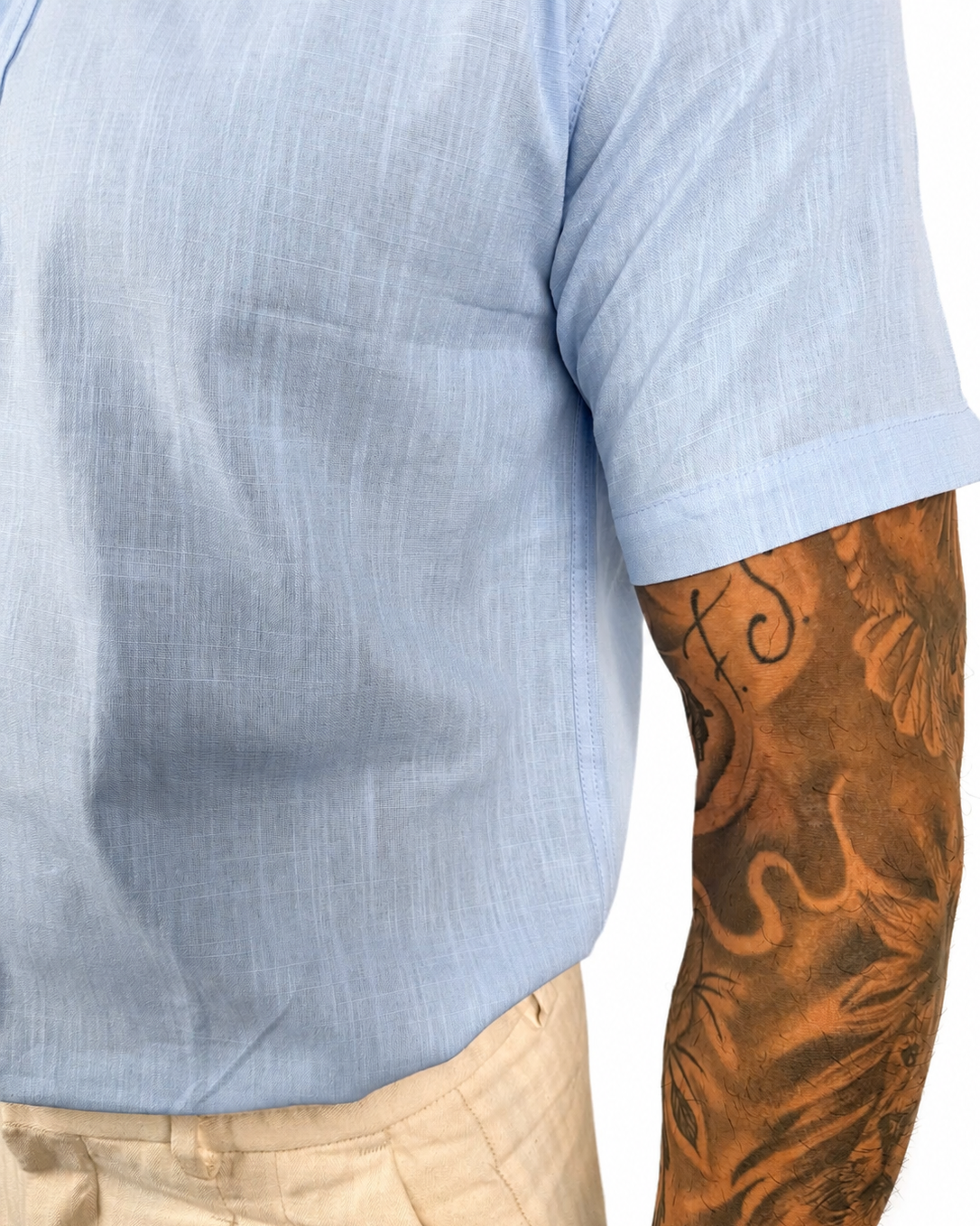 Close-up of a person wearing a light blue short-sleeve linen shirt and beige trousers, with a heavily tattooed arm visible against a plain white background.