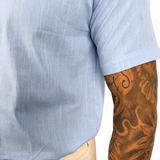 Close-up of a person wearing a light blue short-sleeve linen shirt and beige trousers, with a heavily tattooed arm visible against a plain white background.