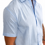 A man wearing a light blue short-sleeve button-down shirt with a textured linen-like fabric, paired with beige pants, shown from the chest down against a white background.